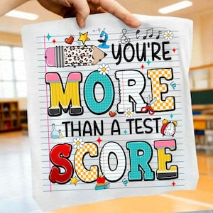 Op de afbeelding: Wit grafisch T-shirt met de tekst "You're More Than a Test Score" in kleurrijke blokletters. Het ontwerp bevat een potlood, muzieknoten, bloemen en andere schoolthema-illustraties.