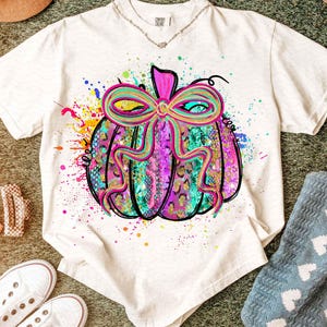 May include: Cream-colored t-shirt featuring a colorful pumpkin design. The pumpkin has a bow and is decorated with a mix of patterns and colors, including pink, teal, and gold. The shirt is part of a fall-themed collection.
