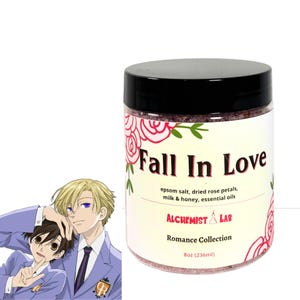 Pode incluir: Um frasco de sais de banho com tampa preta e rótulo que diz "Fall In Love". O rótulo inclui os ingredientes: sal de Epsom, pétalas de rosa secas, leite e mel e óleos essenciais. O frasco faz parte da "Coleção Romance" e contém 236 ml.