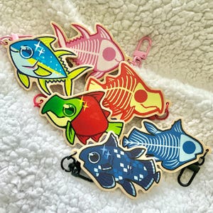 Może przedstawiać: Sześć kolorowych drewnianych breloczków w kształcie ryb z czarnymi metalowymi kółkami. Ryby są pomalowane w różnych stylach, w tym żółta i niebieska ryba z błyszczącymi oczami, czerwona i zielona ryba z błyszczącymi oczami, różowy i biały szkielet ryby, czerwony i żółty szkielet ryby, niebieski i biały szkielet ryby oraz niebieska i biała ryba z błyszczącymi oczami.