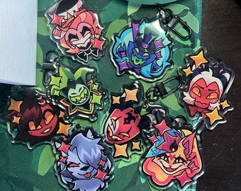 May include: A collection of clear acrylic keychains featuring colorful cartoon character designs. Each keychain has a black metal clasp and is surrounded by star-shaped accents. The characters have various expressions and are in shades of red, blue, green, and orange.