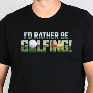 May include: Black t-shirt with the text "I'D RATHER BE GOLFING!" in a colorful golf course design. The shirt features a golf ball and a red flag. The person is wearing blue jeans.