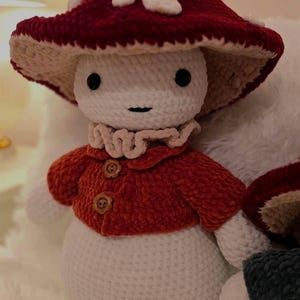 May include: A handmade crochet mushroom doll with a red and white spotted cap, a red jacket with wooden buttons, and a white body. The doll has black eyes and a small smile. The mushroom doll is a whimsical, soft toy.