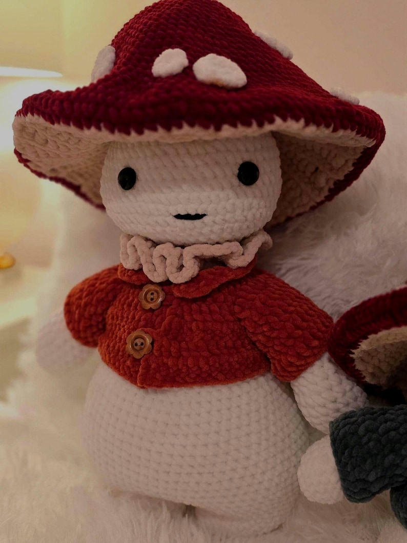 May include: A handmade crochet mushroom doll with a red and white spotted cap, a red jacket with wooden buttons, and a white body. The doll has black eyes and a small smile. The mushroom doll is a whimsical, soft toy.