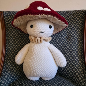 May include: A handmade plush mushroom figure with a white body, a ruffled collar, and a burgundy mushroom cap with white spots. The figure has black eyes and a stitched mouth. The plush is sitting on a patterned chair.