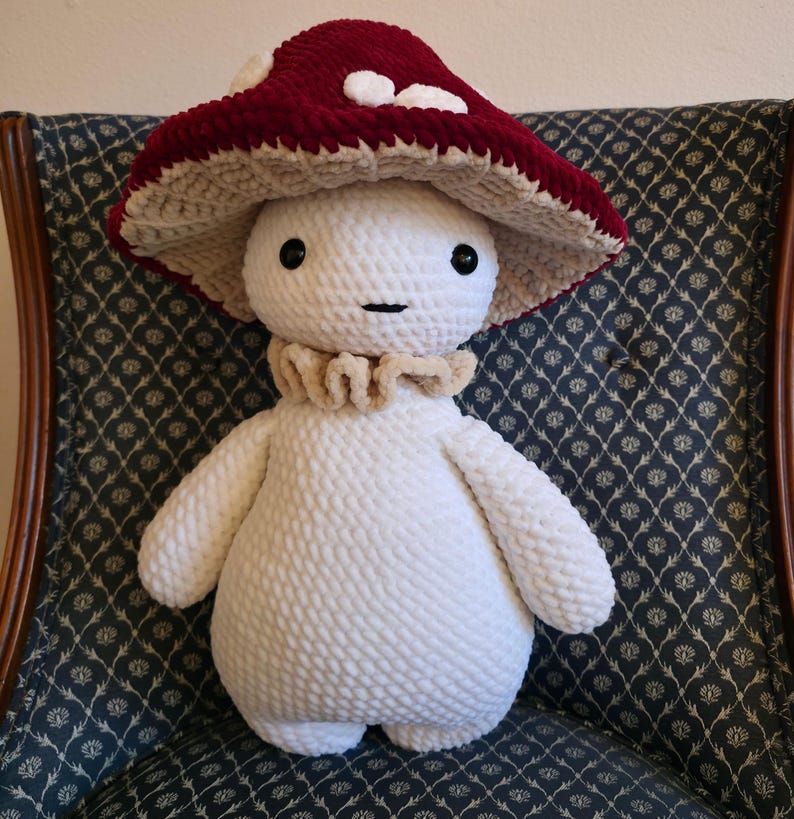 May include: A handmade plush mushroom figure with a white body, a ruffled collar, and a burgundy mushroom cap with white spots. The figure has black eyes and a stitched mouth. The plush is sitting on a patterned chair.
