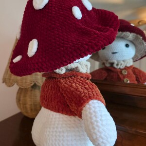 May include: A handmade mushroom-shaped plushie with a red cap featuring white spots. The plushie has a white stem, an orange jacket, and a ruffled collar. The plushie is reflected in a mirror, showcasing its whimsical design.