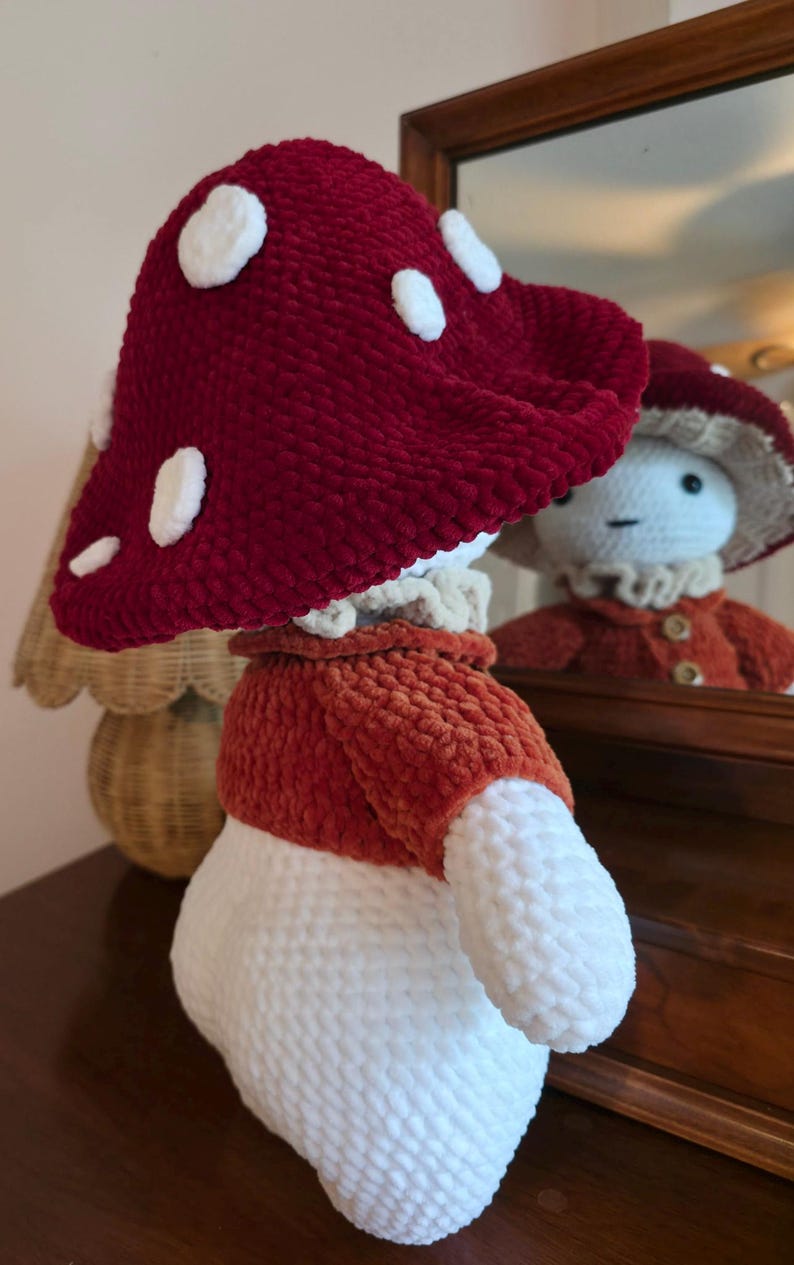 May include: A handmade mushroom-shaped plushie with a red cap featuring white spots. The plushie has a white stem, an orange jacket, and a ruffled collar. The plushie is reflected in a mirror, showcasing its whimsical design.
