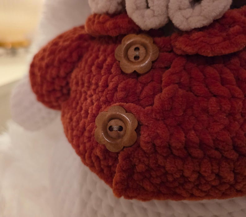 May include: Close-up of a handmade, rust-colored knitted item with two flower-shaped wooden buttons. The item has a textured, plush appearance and is set against a white background. The buttons are a light brown color.