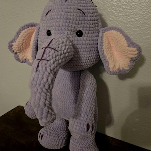 May include: A handmade crochet elephant plush toy made of soft, light purple yarn. The elephant features large ears with light pink inner sections, dark purple stitched details on the trunk and head, and small black plastic eyes.