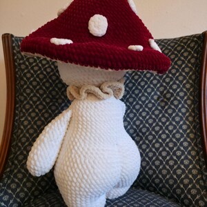 May include: A handmade plush mushroom toy with a burgundy cap and white spots. The stem and arms are white, and the mushroom has a ruffled collar. The toy is made of a soft, textured material.