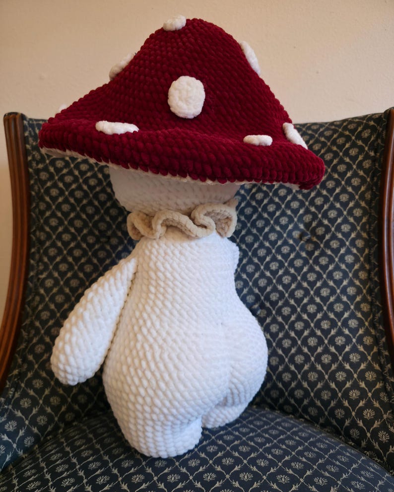May include: A handmade plush mushroom toy with a burgundy cap and white spots. The stem and arms are white, and the mushroom has a ruffled collar. The toy is made of a soft, textured material.