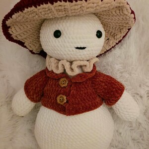 May include: A plush mushroom figure, approximately 14 inches tall, with a burgundy and cream-colored mushroom cap. It has a white body, a rust-colored jacket with flower-shaped buttons, and a ruffled collar. The figure has a smiling face.