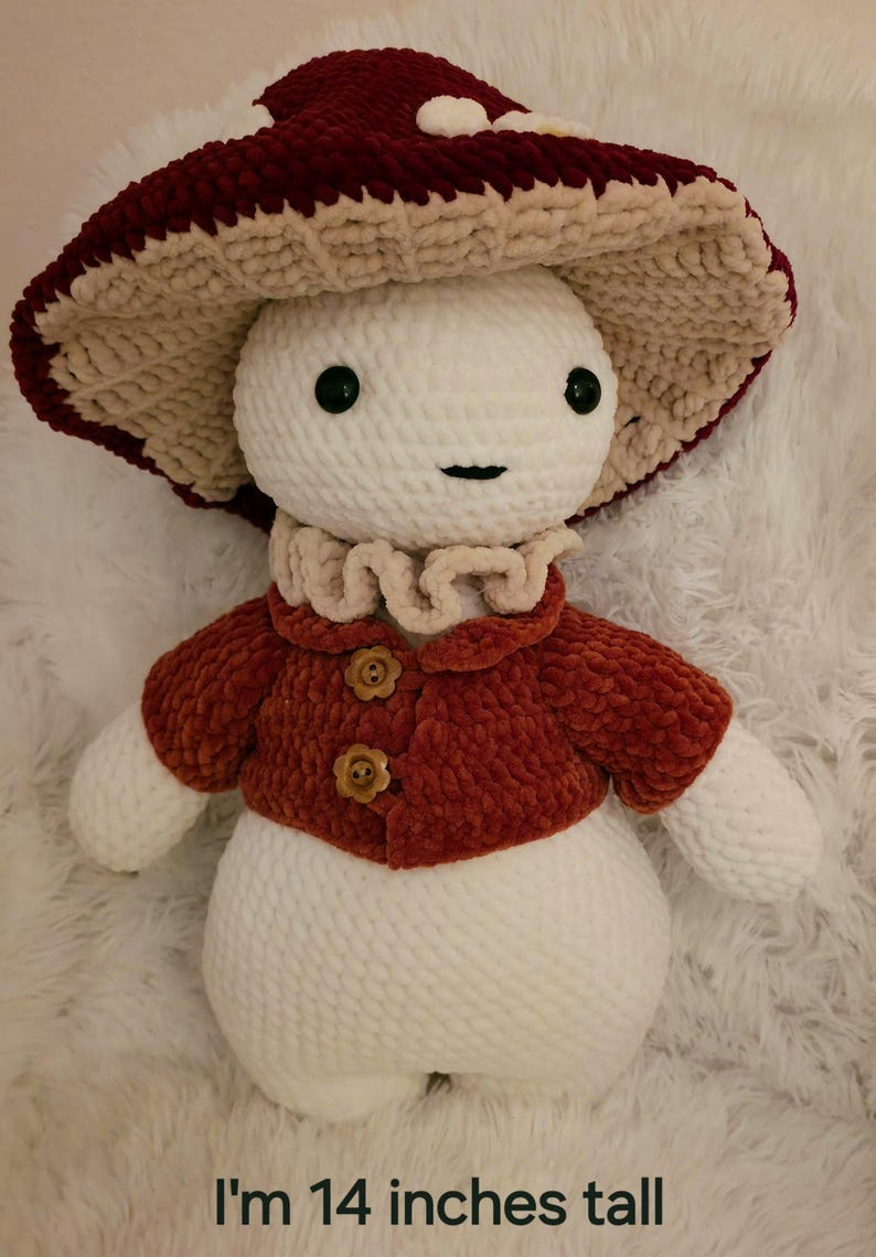May include: A plush mushroom figure, approximately 14 inches tall, with a burgundy and cream-colored mushroom cap. It has a white body, a rust-colored jacket with flower-shaped buttons, and a ruffled collar. The figure has a smiling face.