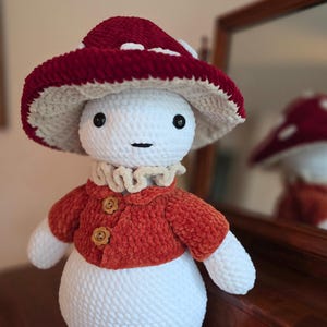 May include: A handmade crochet mushroom doll with a red and white mushroom cap, a rust-colored jacket with wooden buttons, and a ruffled collar. The doll has a white body and arms, and black button eyes.