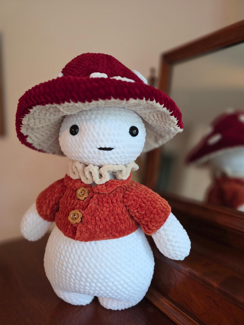 May include: A handmade crochet mushroom doll with a red and white mushroom cap, a rust-colored jacket with wooden buttons, and a ruffled collar. The doll has a white body and arms, and black button eyes.