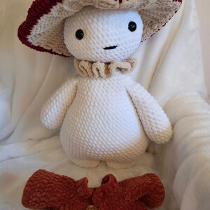 May include: A handmade crochet mushroom plush toy with a burgundy and cream-colored mushroom cap. The plush has a white body, a beige collar, and a matching burgundy jacket with wooden buttons. The toy is soft and cuddly.