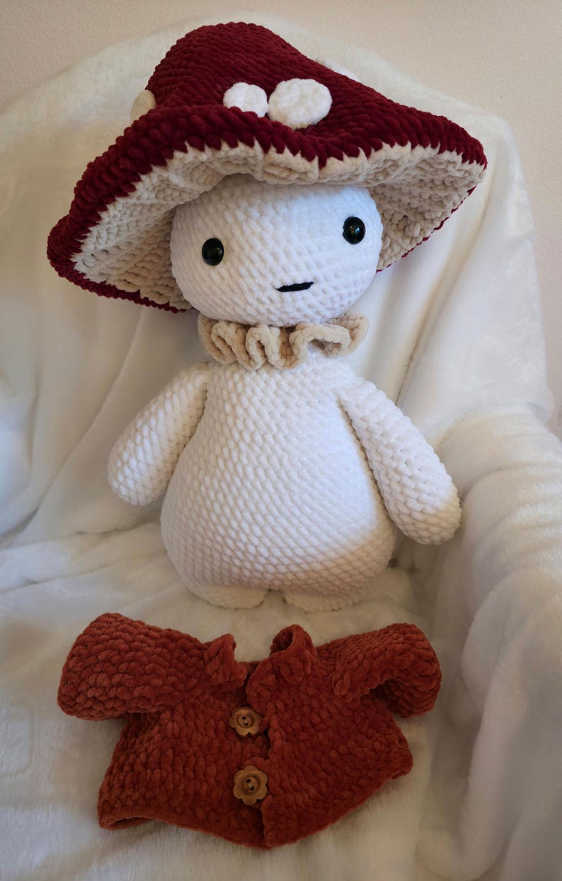 May include: A handmade crochet mushroom plush toy with a burgundy and cream-colored mushroom cap. The plush has a white body, a beige collar, and a matching burgundy jacket with wooden buttons. The toy is soft and cuddly.