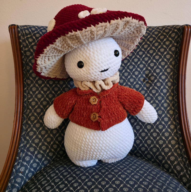 May include: A handmade plush mushroom figure with a red and cream-colored mushroom cap, a white body, and a red jacket with wooden buttons. The figure has a smiling face and is sitting on a patterned chair.
