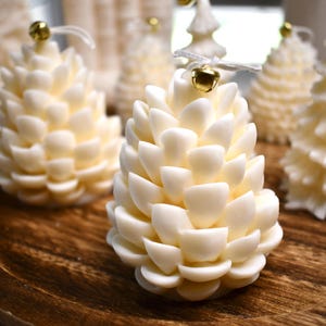 Puede incluir: Velas con forma de piña de color crema con campanas doradas unidas por una cuerda blanca. Las velas están dispuestas sobre una bandeja de madera. También se ven otras velas en el fondo. Decoración festiva.