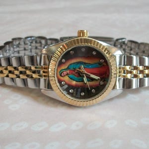 May include: A silver and gold wristwatch with a two-tone metal band. The watch face features a colorful image of the Virgin Mary.