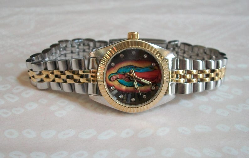 May include: A silver and gold wristwatch with a two-tone metal band. The watch face features a colorful image of the Virgin Mary.