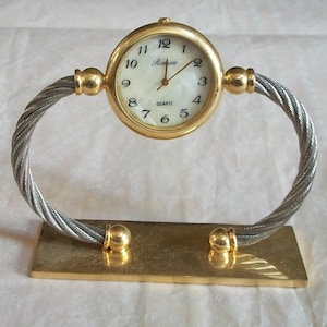 May include: A gold-toned quartz desk clock with a white face and black numerals. The clock is supported by a twisted silver-tone metal frame and a rectangular gold-tone base. The clock face reads "Ronica".