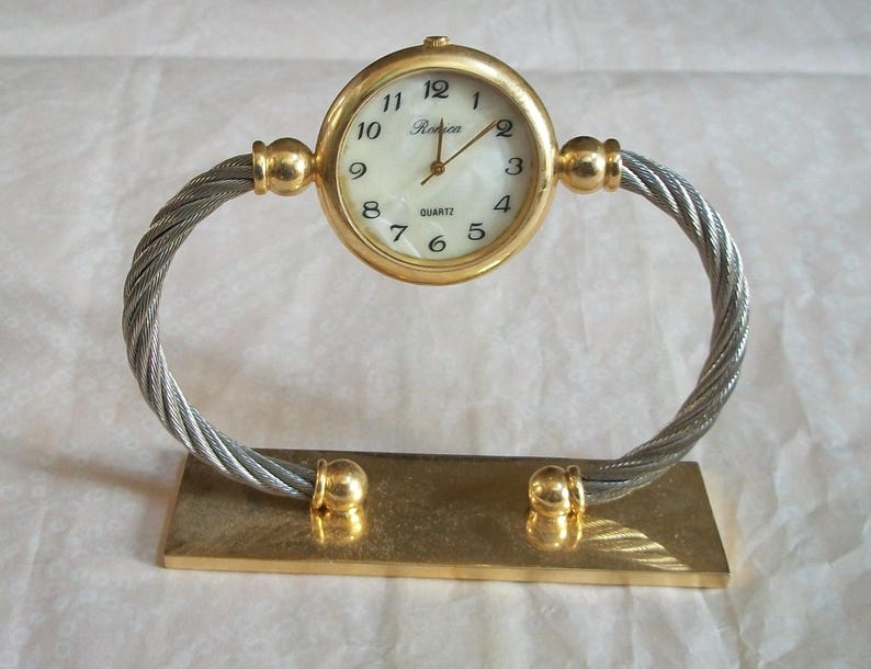 May include: A gold-toned quartz desk clock with a white face and black numerals. The clock is supported by a twisted silver-tone metal frame and a rectangular gold-tone base. The clock face reads "Ronica".