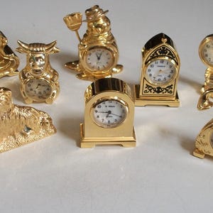 May include: A collection of gold-colored miniature clocks in various shapes, including a cat, bull, and eagle. Each clock has a white face with black numerals and hands. The clocks are displayed on a white surface.