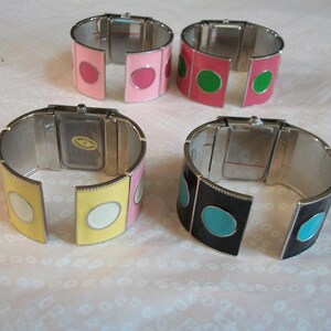 May include: Four silver bangle watches with different colored enamel accents. The watches have white faces and are open to show the watch mechanism. The colors of the enamel accents are pink, green, yellow, and black.