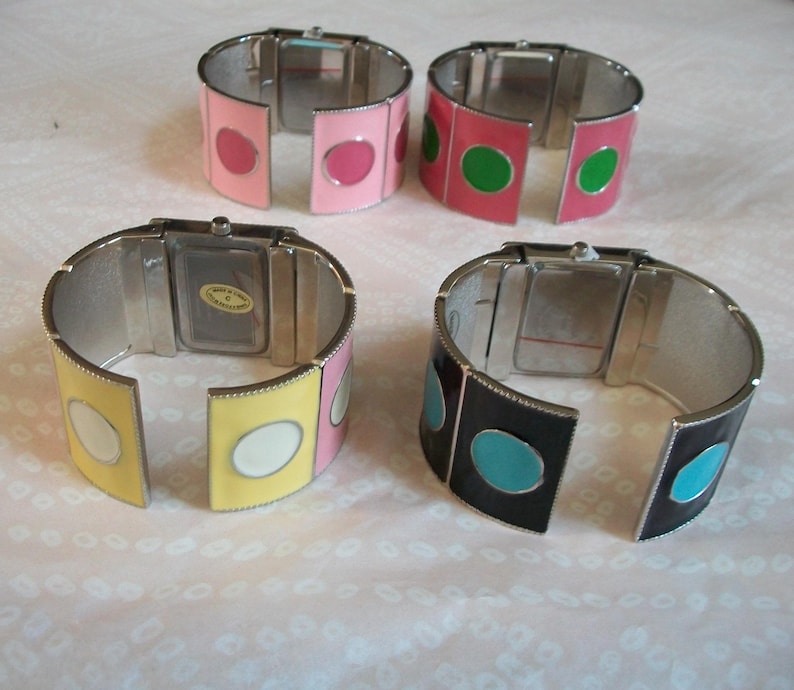 May include: Four silver bangle watches with different colored enamel accents. The watches have white faces and are open to show the watch mechanism. The colors of the enamel accents are pink, green, yellow, and black.