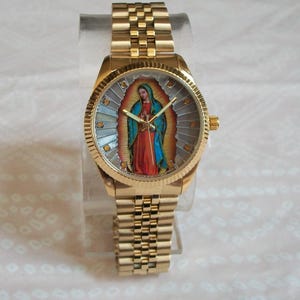 May include: A gold-tone wristwatch with a round face featuring a colorful image of the Virgin Mary. The watch has a gold-tone metal band.