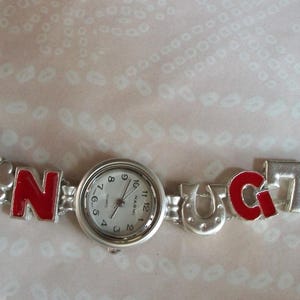 May include: A silver charm bracelet watch with red enamel letters spelling "B.A.T.N.O.C.T.O.". The watch face is white with black numbers and hands.