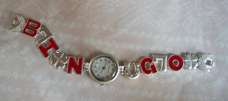 May include: A silver charm bracelet watch with red enamel letters spelling "B.A.T.N.O.C.T.O.". The watch face is white with black numbers and hands.