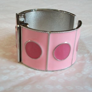 May include: A silver bangle bracelet with a pink enamel design featuring three large circles. The bracelet is open and ready to be worn.