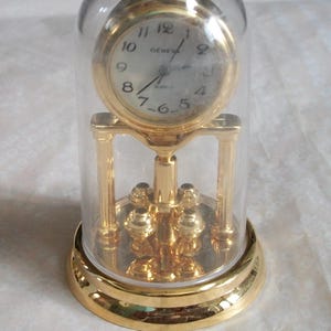 May include: A gold-toned quartz clock encased in a clear glass dome. The clock face is white with black numerals and hands, and the word "GENEVA" is printed above the center. The clock sits on a decorative gold base.
