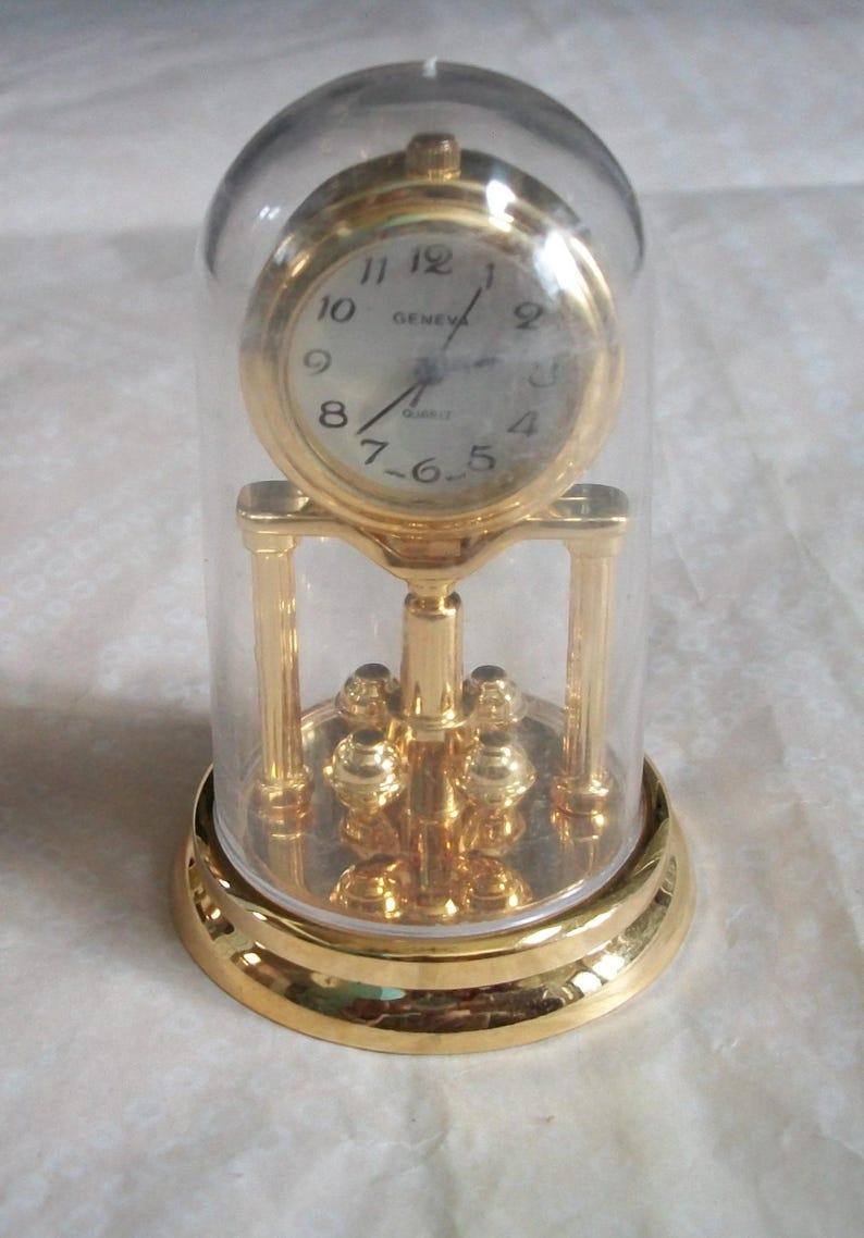 May include: A gold-toned quartz clock encased in a clear glass dome. The clock face is white with black numerals and hands, and the word "GENEVA" is printed above the center. The clock sits on a decorative gold base.