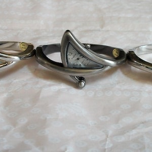 May include: Three silver-toned bangle watches with triangular faces and small, round accents. The watch faces are white with black numerals. The bracelets are displayed on a light pink patterned surface.