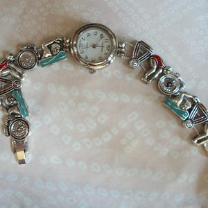 May include: A silver watch with a white face and black numbers. The watch band is made of silver and has charms of swimmers and lifeguards.