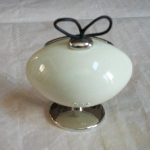 May include: Cream-colored, egg-shaped container with a silver base and a black loop handle. The container has a glossy finish and a silver rim. The base is circular and made of silver metal.