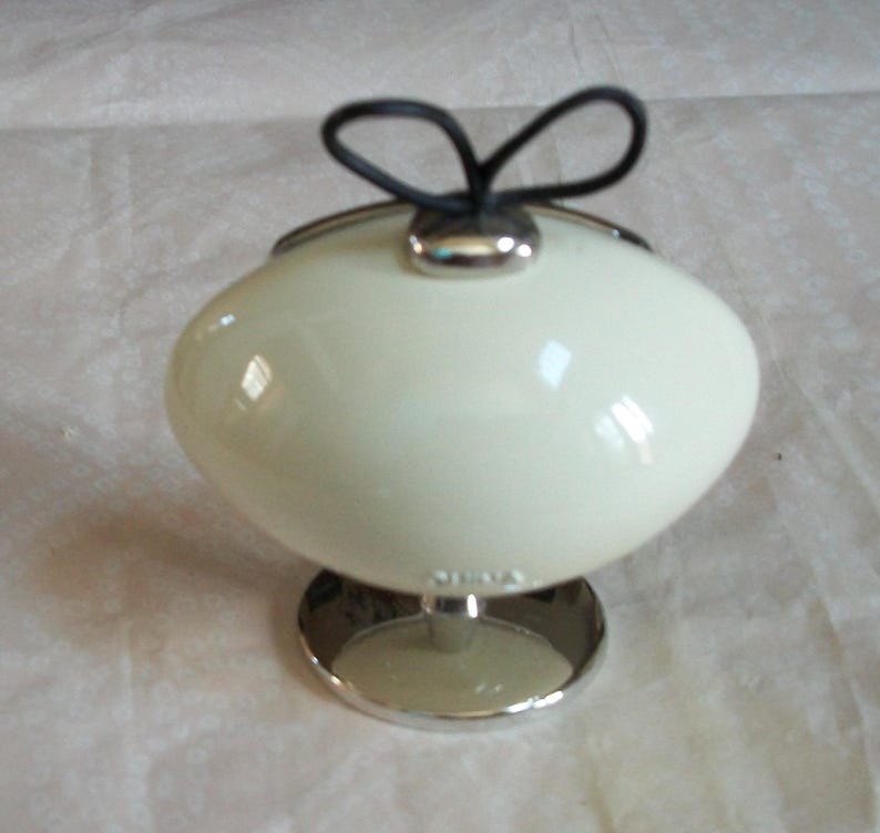 May include: Cream-colored, egg-shaped container with a silver base and a black loop handle. The container has a glossy finish and a silver rim. The base is circular and made of silver metal.