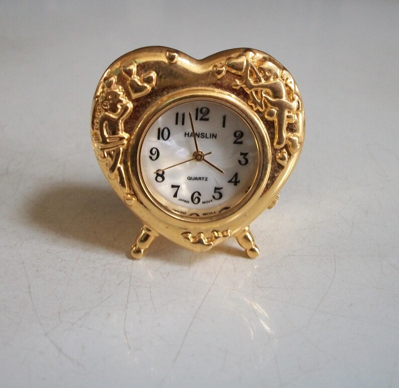 May include: A gold-colored heart-shaped quartz clock with a white face and black numerals. The clock has decorative cherub figures on the sides and stands on three small feet. The clock face reads "Hanslin".