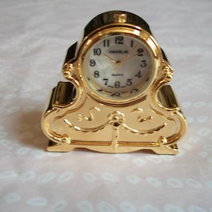 May include: A gold-colored quartz clock with an oval face and Roman numerals. The clock has a decorative, ornate design and the brand name "HANSLIN" is visible on the clock face.