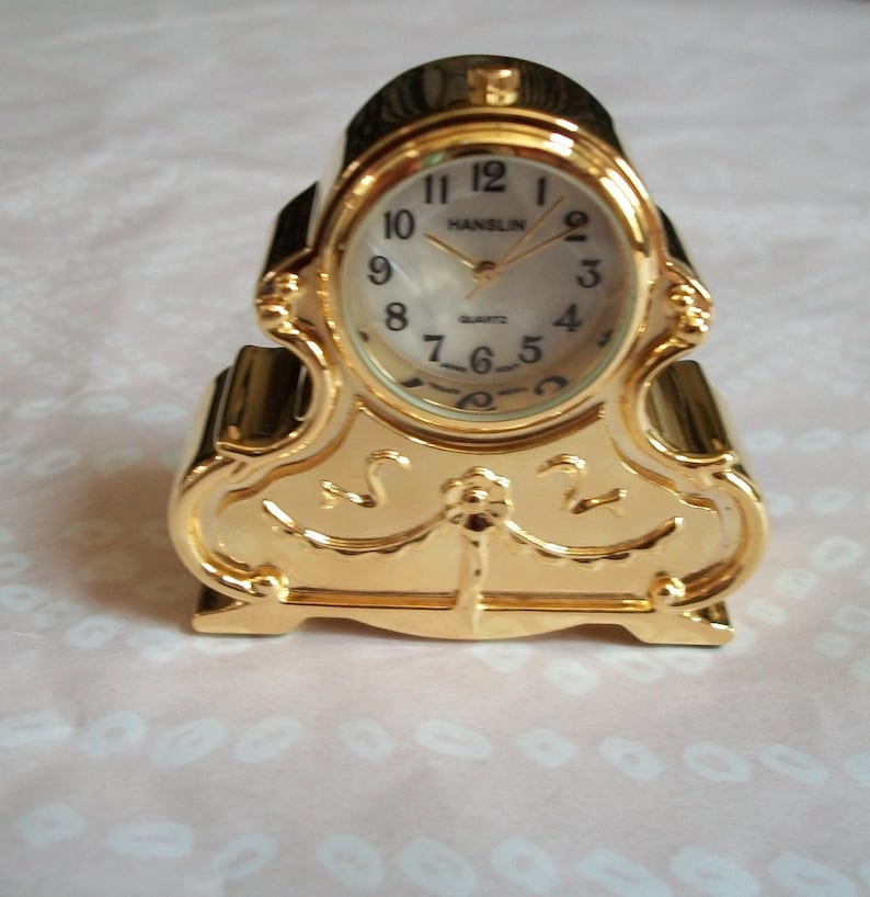 May include: A gold-colored quartz clock with an oval face and Roman numerals. The clock has a decorative, ornate design and the brand name "HANSLIN" is visible on the clock face.