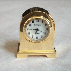 May include: A gold-colored quartz clock with a white face and black numerals. The clock has a rectangular base and an arched top. The clock face reads "St. Marin" and "Quartz".