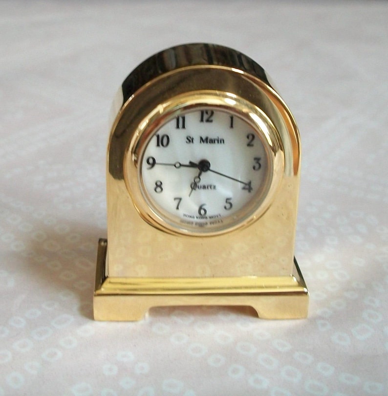 May include: A gold-colored quartz clock with a white face and black numerals. The clock has a rectangular base and an arched top. The clock face reads "St. Marin" and "Quartz".