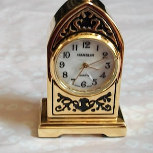 May include: A gold-colored quartz clock with a decorative, arched top and base. The clock face is white with black numerals and hands. The brand name "HANSLIN" is printed on the clock face. The clock has an ornate design.