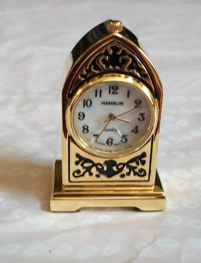 May include: A gold-colored quartz clock with a decorative, arched top and base. The clock face is white with black numerals and hands. The brand name "HANSLIN" is printed on the clock face. The clock has an ornate design.