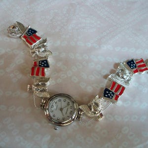 May include: A silver watch bracelet with American flag charms. The watch face is white with black numbers and hands.