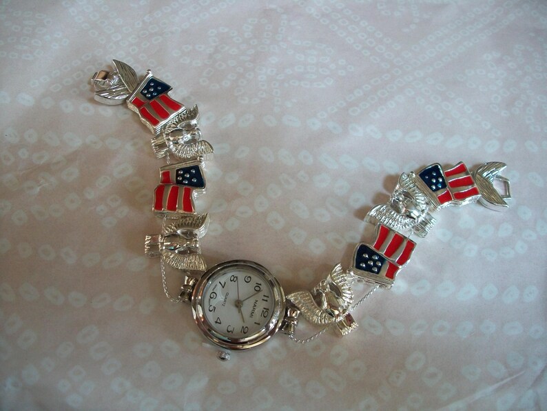 May include: A silver watch bracelet with American flag charms. The watch face is white with black numbers and hands.